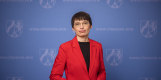 Ministerin Josefine Paul steht vor einer blauen Pressewand und schaut mit einem leichten Lächeln in die Kamera. Der Oberkörper steht leicht schräg nach rechts.